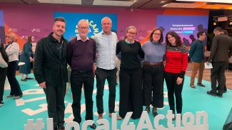 Six people smiling at a convention, standing in front of a sign reading '#Local4Action'